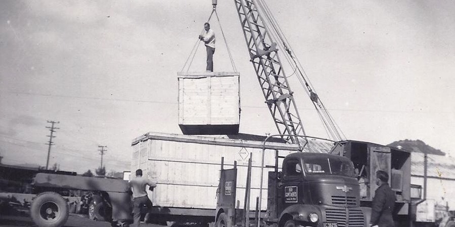1940s B&W photo of small truck crane lifting crate onto transport trailer, with worker standing on crate